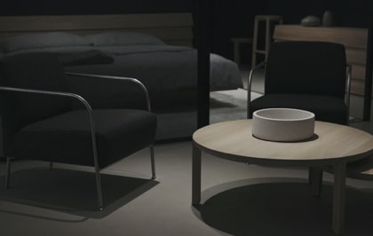 A dimly-lit room featuring two black, deep-seated, cushioned armchairs with short backs, around a circular coffee table with a cylindrical bowl placed in the center