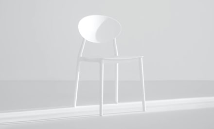 A white-tinctured, washed-out view of a small, white plastic chair of minimilistic design with an ovular back, highlighted by a stream of sunlight shining from the right side